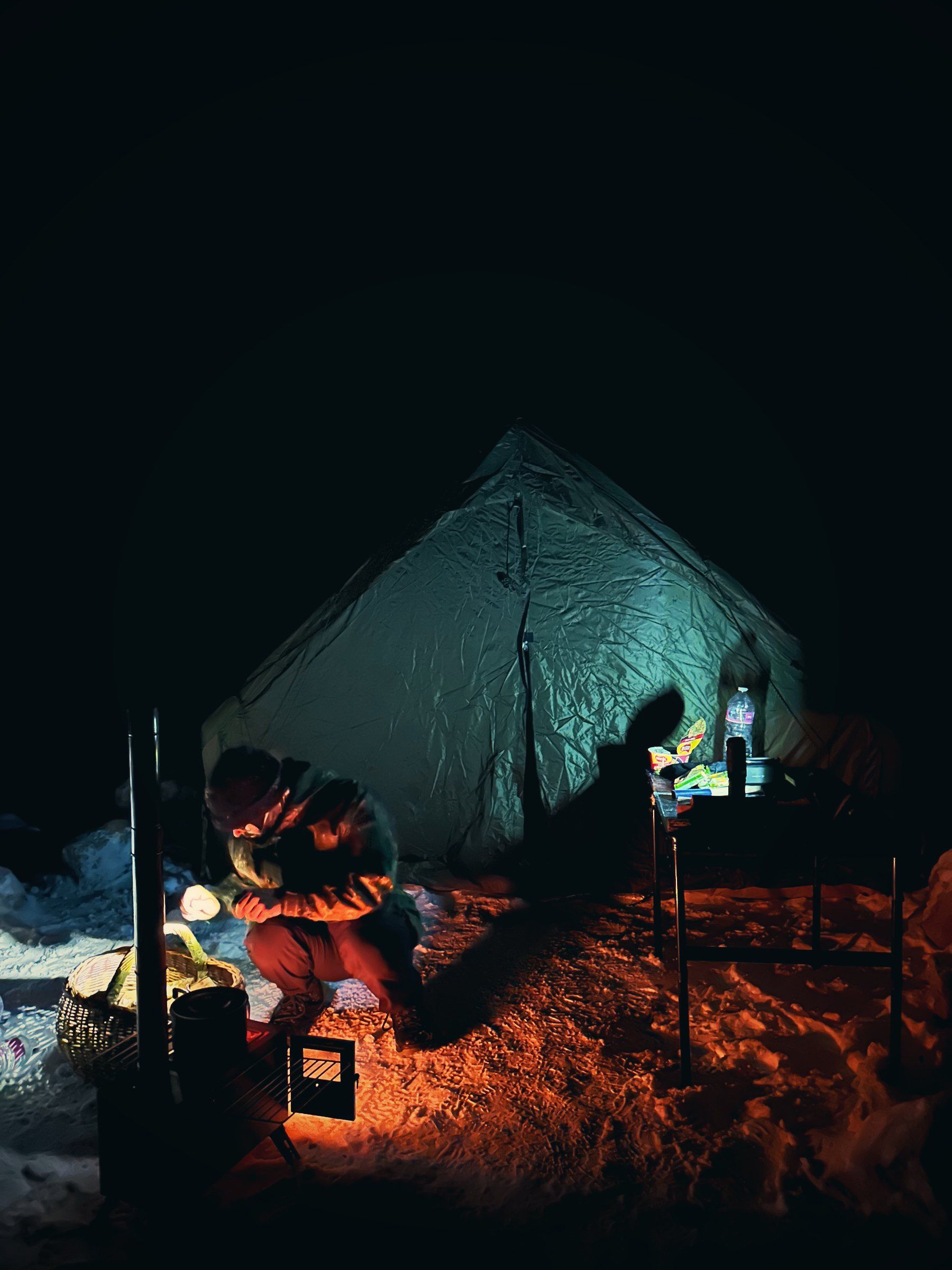Beleuchtetes Zelt im Schnee in der Nacht, davor der glühende Ofen