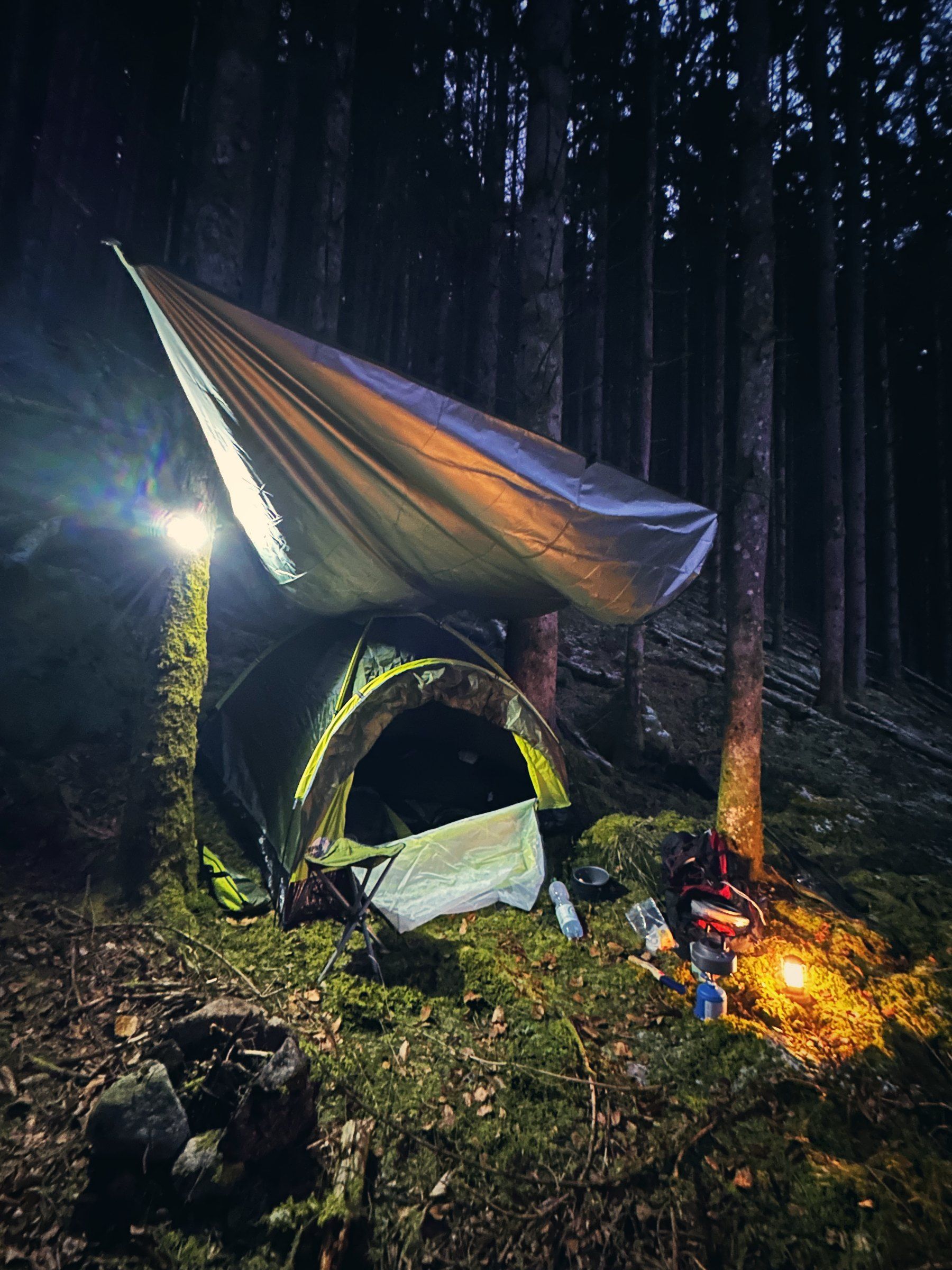 Zelt unter einem Tarp im dunklen Winterwald, daneben eine Laterne